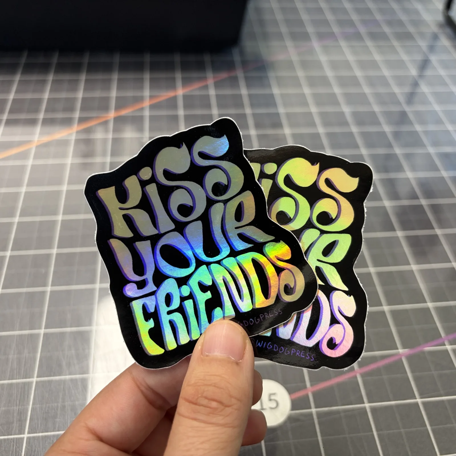 Two Kiss Your Friends holographic stickers held over a cutting mat