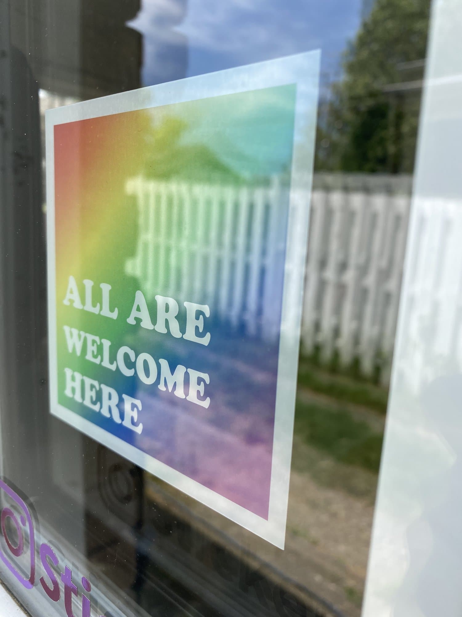 An 'All Are Welcome Here' rainbow window cling on a storefront door