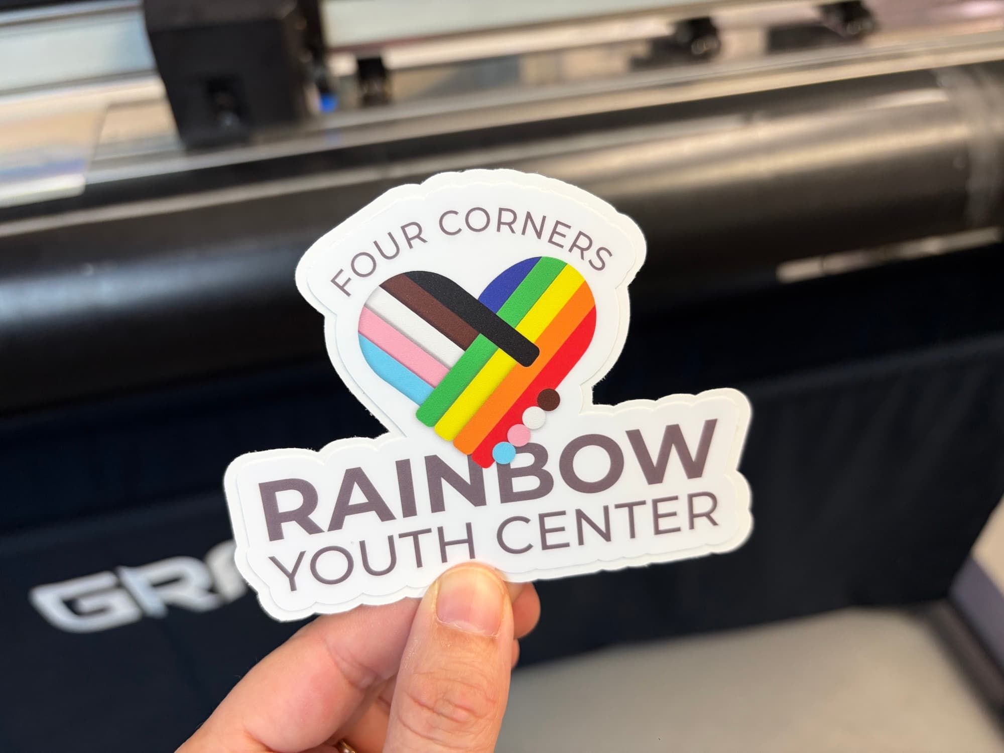 Four Corners Rainbow Youth Center die cut sticker with a progress Pride heart, held in front of the cutter