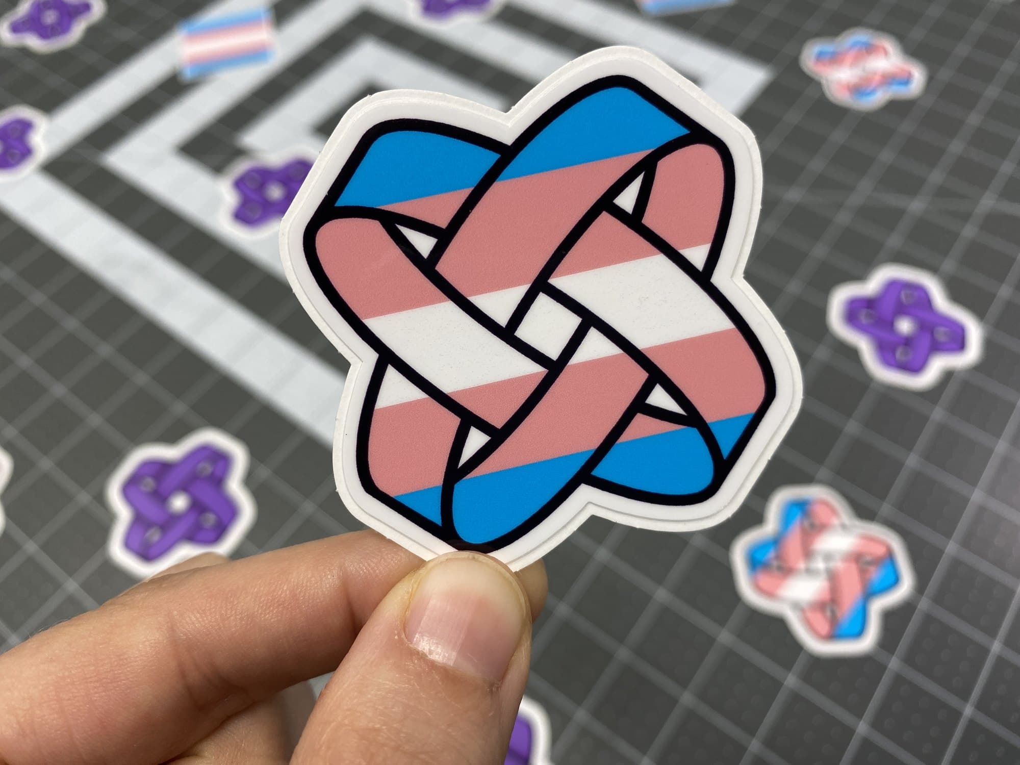 Genderbands trans flag knot sticker held over a sheet of freshly cut stickers