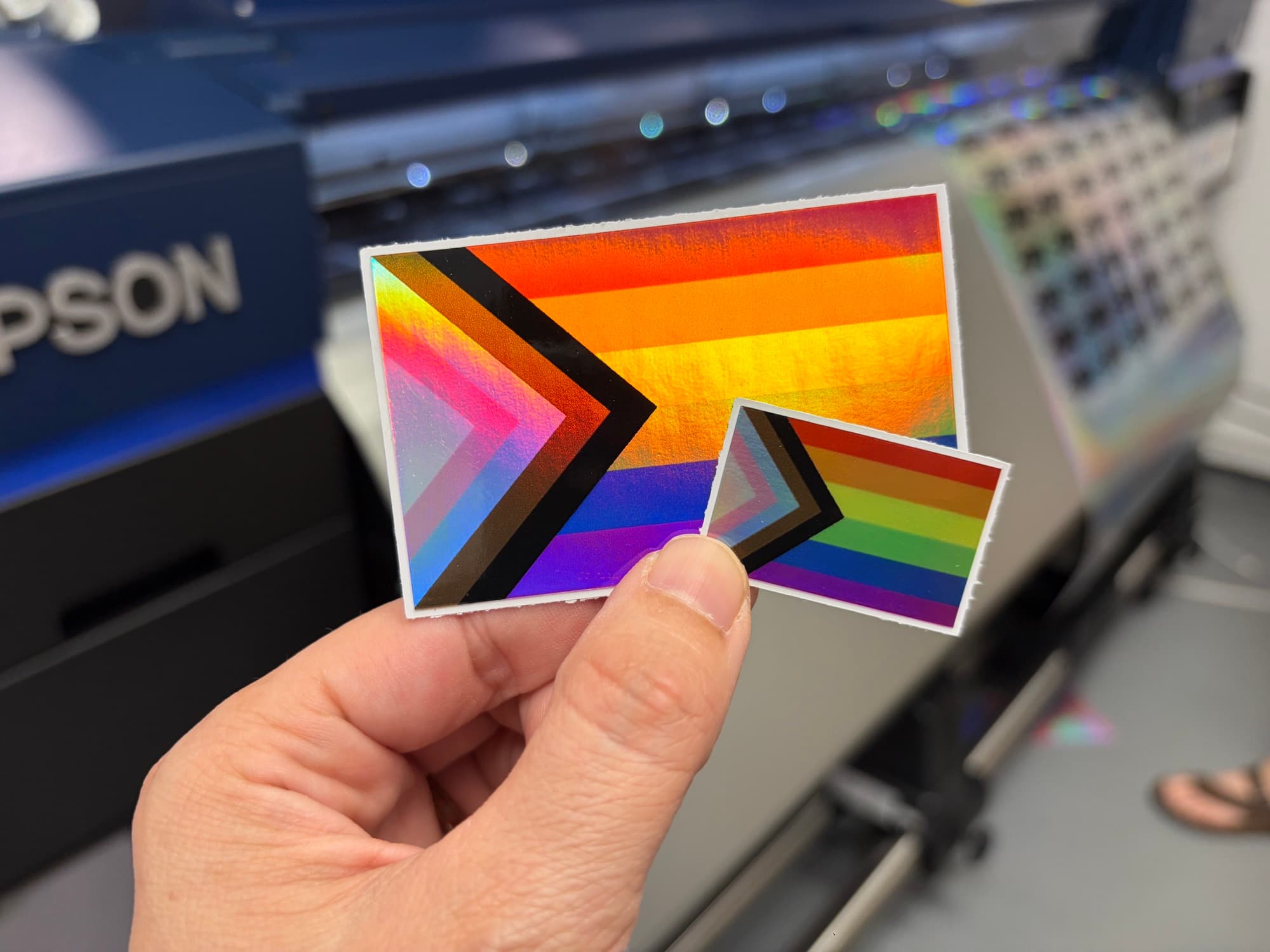 Two holographic progress Pride flag stickers held up in front of the printer