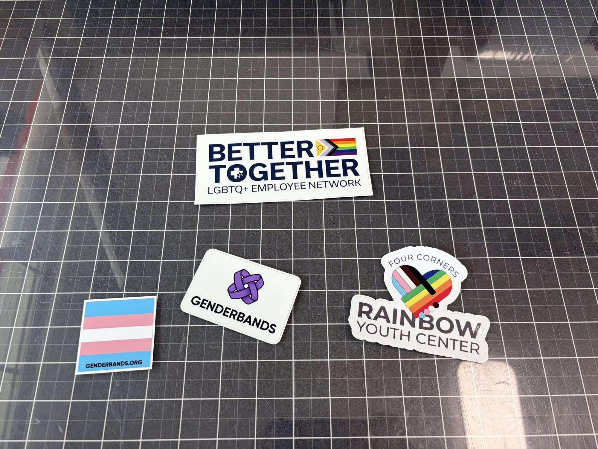 Stickers for an LGBTQ+ employee network, Genderbands, and Four Corners Rainbow Youth Center on the cutting mat
